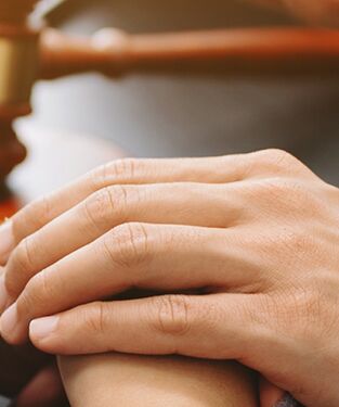 Nahaufnahme zweier Hände, die sich beruhigend berühren, im Hintergrund ist ein Richterhammer zu sehen.