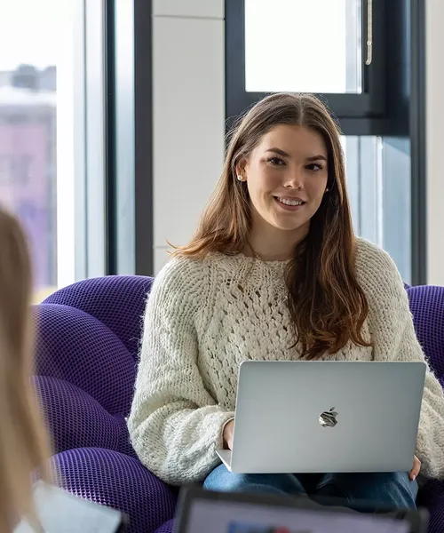 Junge Frau sitzt auf einem lila Sessel mit Laptop auf dem Schoß und lächelt eine andere Person im Vordergrund an.