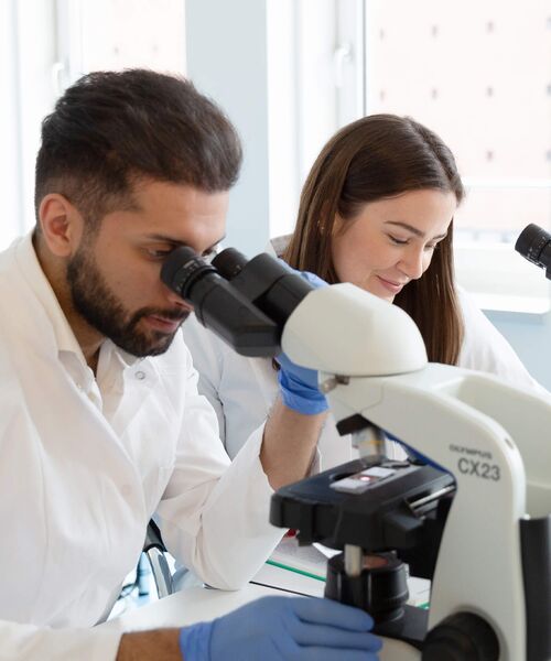 Zwei Studierende im weißen Kittel arbeiten im Labor, einer schaut durch ein Mikroskop, die andere macht sich Notizen.