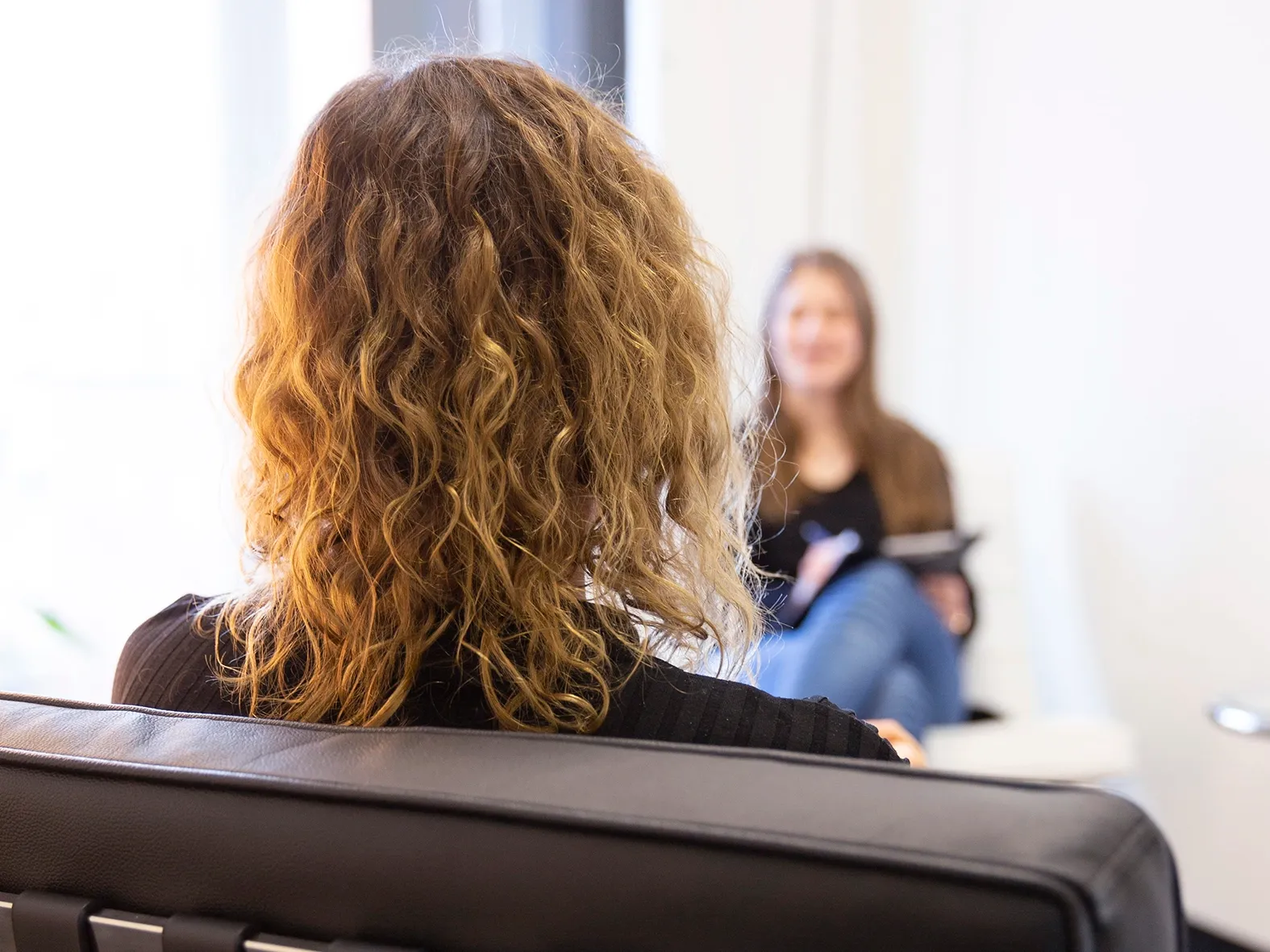 Zwei Personen in einem Gespräch, im Vordergrund eine Person mit lockigen Haaren, im Hintergrund eine sitzende Person mit Notizblock in heller Umgebung.
