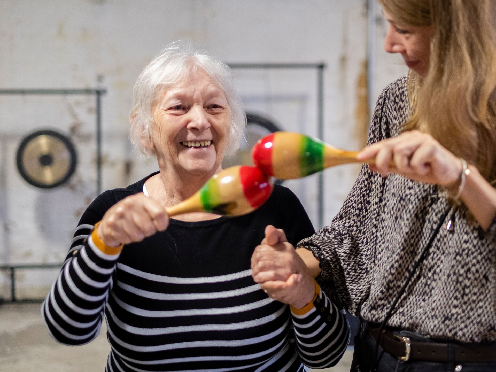 Ältere Frau lächelt und schüttelt bunte Maracas, während sie von einer anderen Person unterstützt wird.