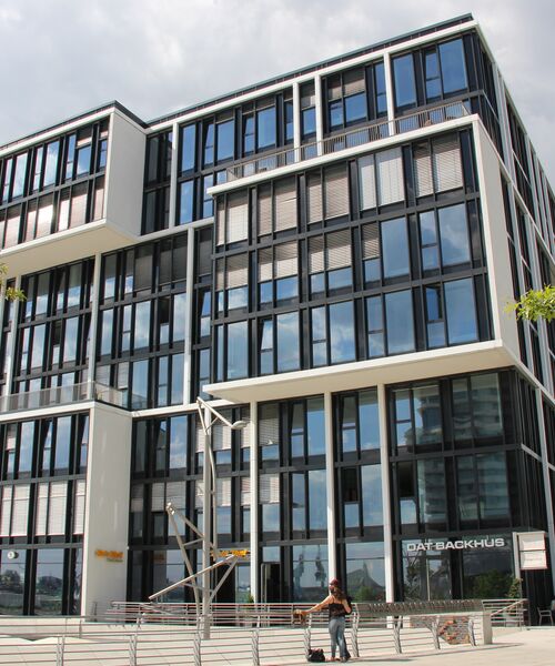 Modernes Gebäude mit großflächiger Glasfassade und markanten Vorsprüngen, im Erdgeschoss ein Café und Passanten davor.