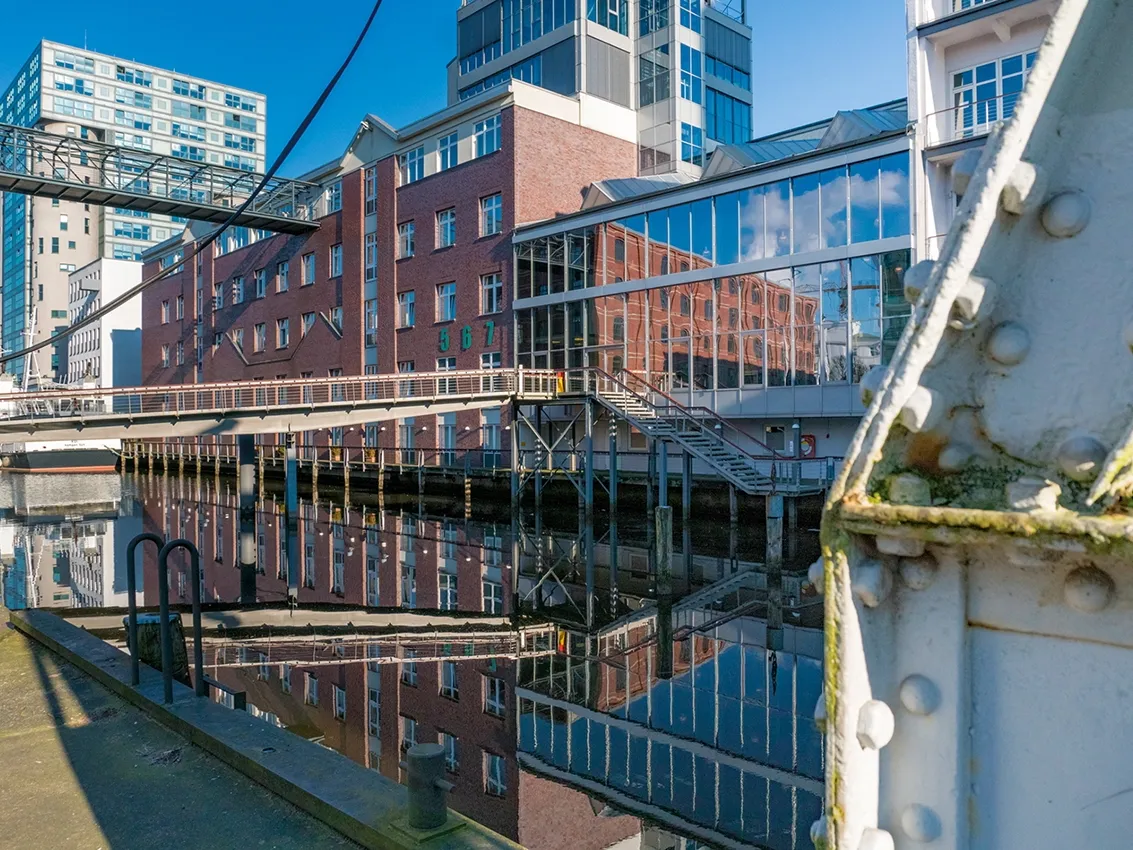 Moderne Gebäudefront am Wasser mit Backstein- und Glasfassaden, deren Spiegelung sich klar in der ruhigen Wasseroberfläche zeigt.