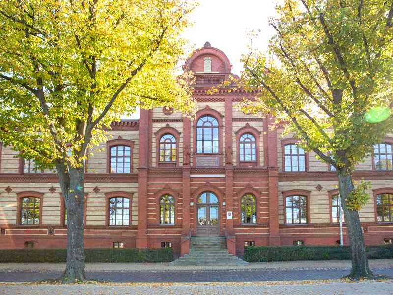 Historisches Backsteingebäude mit symmetrischer Fassade und Rundbogenfenstern, umgeben von Bäumen in herbstlicher Färbung.