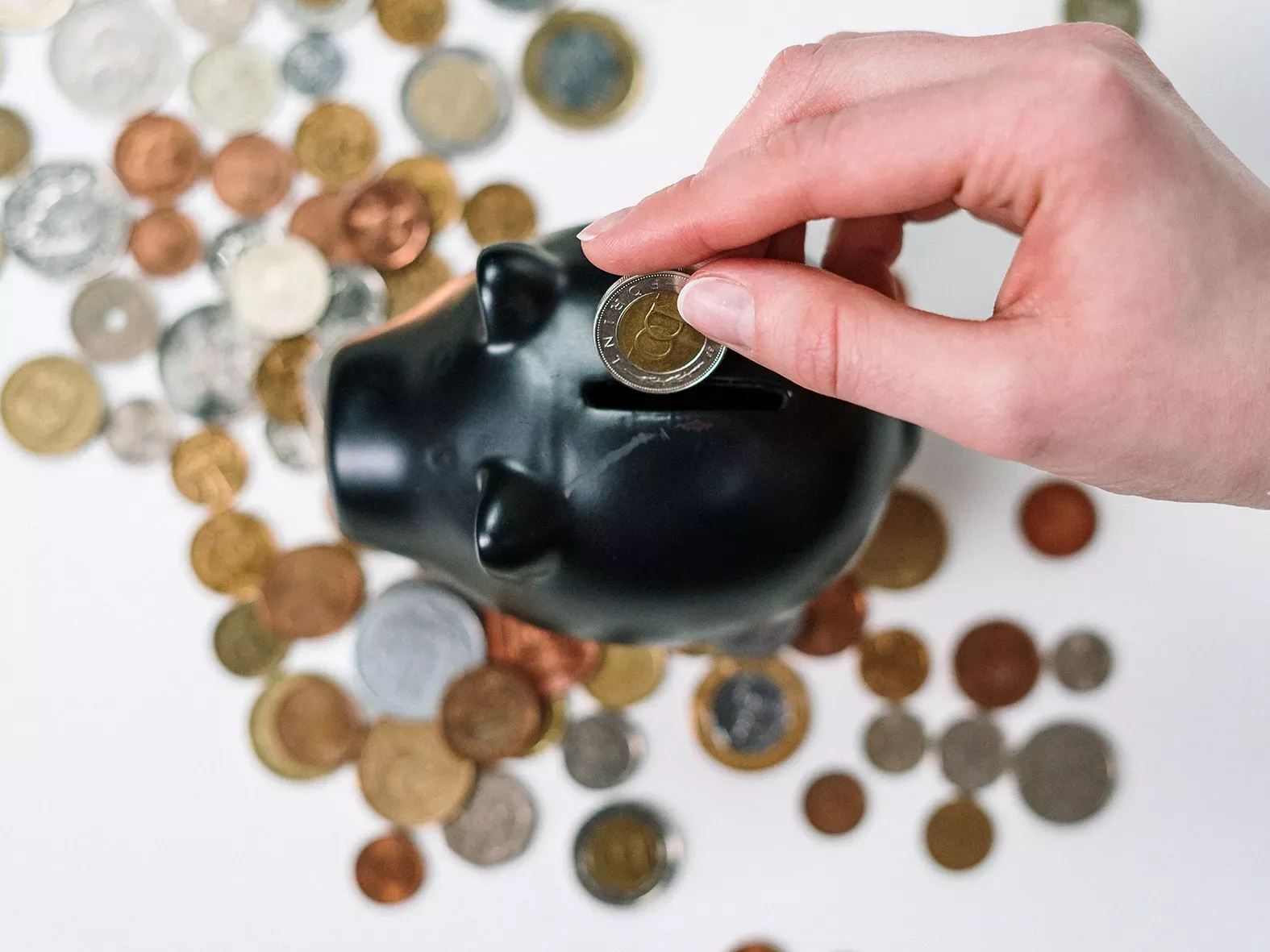 Hand wirft eine Münze in ein schwarzes Sparschwein, umgeben von weiteren Münzen auf dem Tisch.