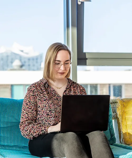 Frau mit Brille sitzt auf einem Sofa und arbeitet konzentriert an einem Laptop.
