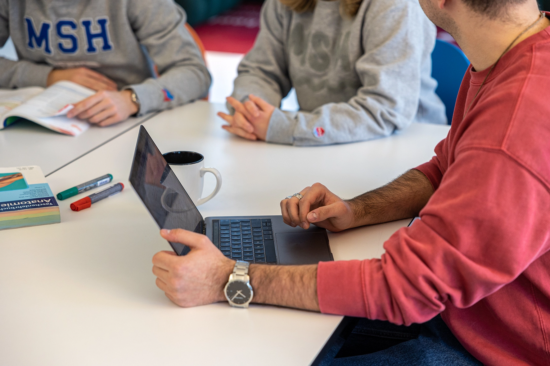 Studierende arbeiten gemeinsam an einem Tisch mit Laptop, Tasse, Stiften und einem Anatomiebuch.