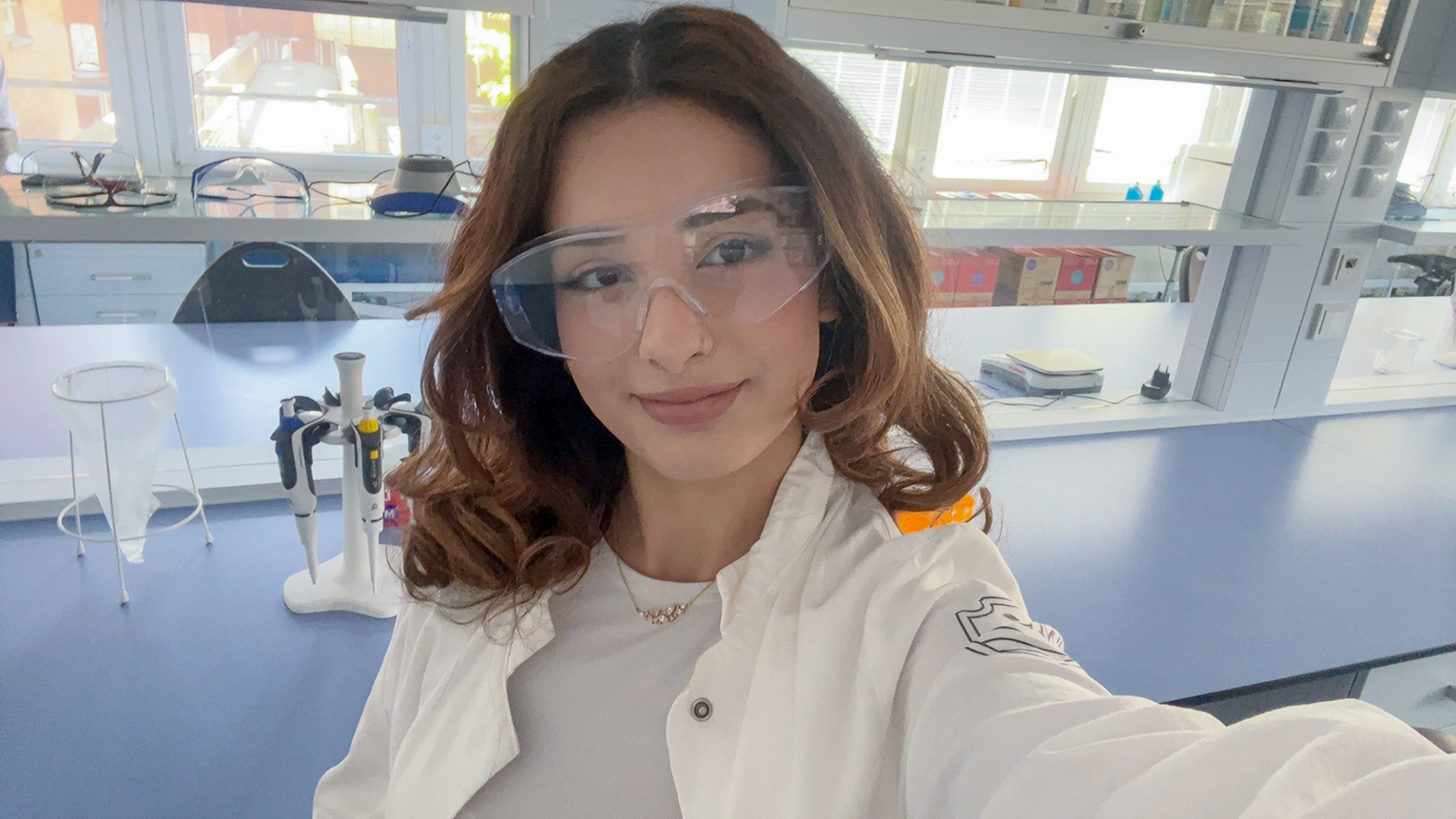 Eine junge Frau im weißen Laborkittel und Schutzbrille macht ein Selfie in einem modernen Labor mit wissenschaftlichen Geräten im Hintergrund.